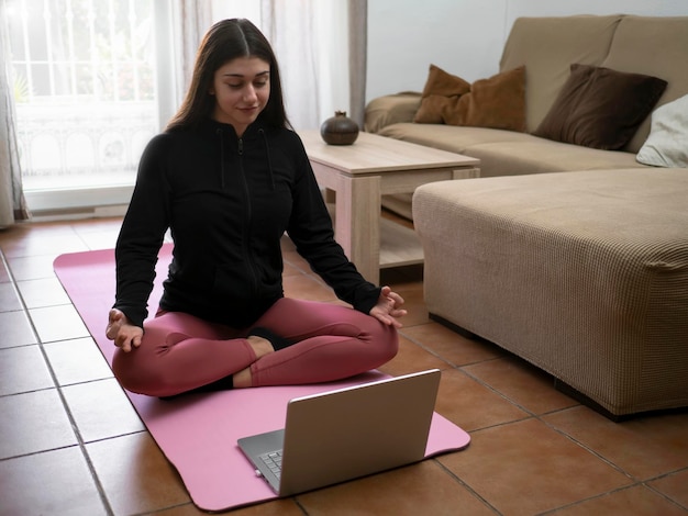 Quiet girl meditating in front of laptop at home sitting on the mat in the living room