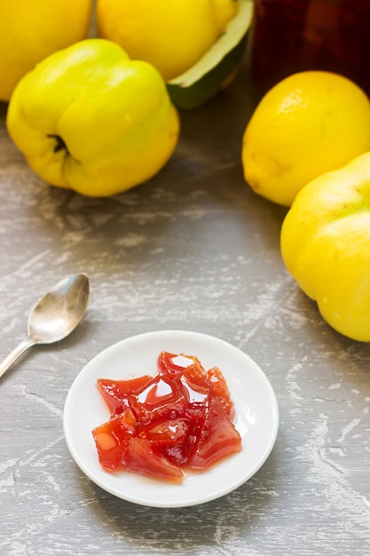 Marmellata di mele cotogne su un piattino bianco, frutti di mela cotogna e vasetti di marmellata su uno sfondo grigio.