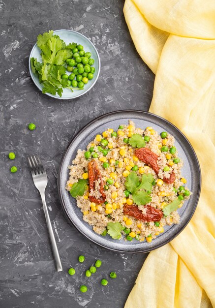 Каша из киноа с зеленым горошком, кукурузой и сушеными помидорами на керамической тарелке на серой бетонной поверхности и желтой ткани