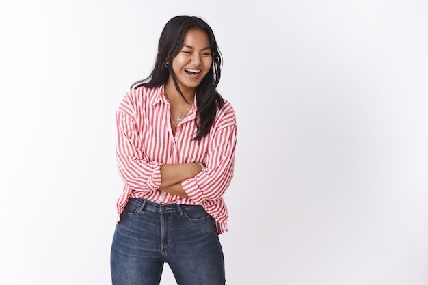 Quit joking. Portrait of happy and carefree joyful cute asian girl having fun laughing from funny friend having great sense of humour looking right cross hands over chest against white wall