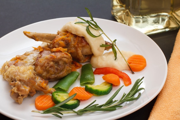 Ножки кролика, запеченные в белом вине с соусом бешамель на керамической тарелке с овощами и розмарином. Диетическое мясо кролика, приготовленное в духовке.