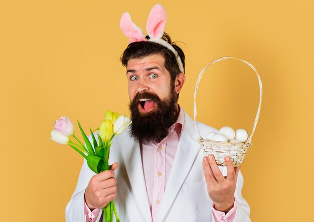 Uomo coniglio in orecchie da coniglio con fiori e uova nel cestino. concetto di celebrazione di pasqua. uomo barbuto in abito con fiore di primavera.