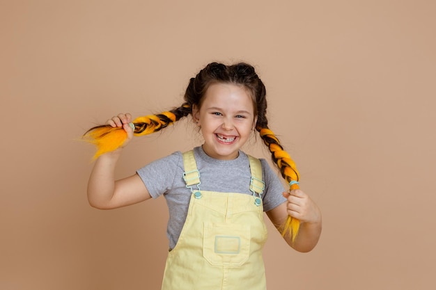 Сияющая довольная молодая девочка с желтыми косами из канекалона смеется и дергает за косички, смотрит в камеру, улыбается в желтом комбинезоне и серой футболке на бежевом фоне