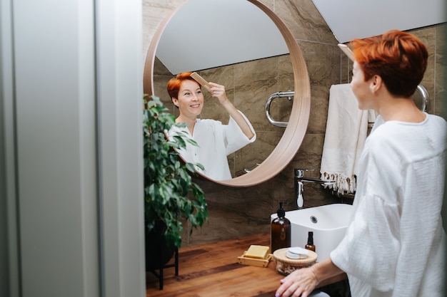 Donna matura dai capelli corti radiante che si pettina i capelli con un pettine di legno. è in piedi davanti allo specchio in un bagno, riflesso in un fuoco.