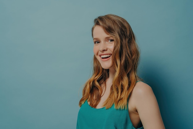 Radiant young woman with wavy hair and green top laughing joyfully against a cool blue background