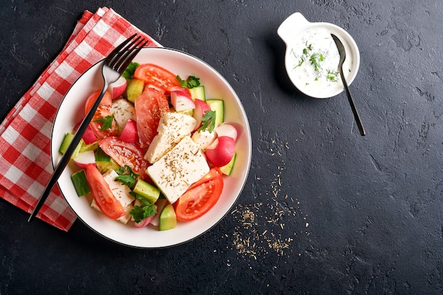 Radish, cucumber, tomato, pepper and feta cheese with spices pepper and olive oil in white bowl on black slate, stone or concrete background. Healthy food concept. Top view.