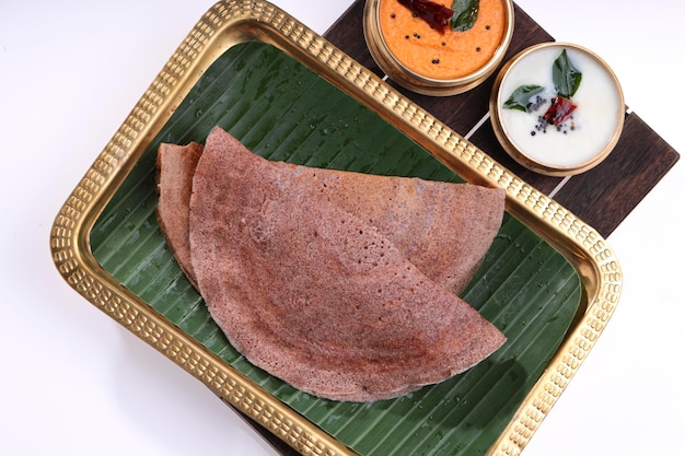 Ragi Dosa, healthy south Indian breakfast item arranged on a rectangle brass plate lined with banana leaf and coconut chutneys placed beside it.