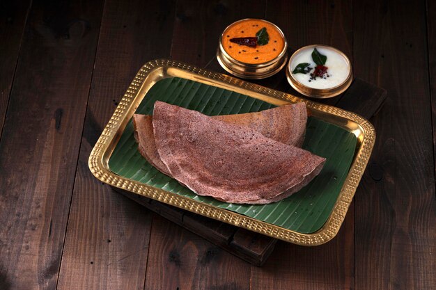 Ragi Dosa, healthy south Indian breakfast item arranged on a rectangle brass plate lined with banana leaf and coconut chutneys placed beside it.