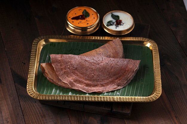Ragi Dosa, healthy south Indian breakfast item arranged on a rectangle brass plate lined with banana leaf and coconut chutneys placed beside it.
