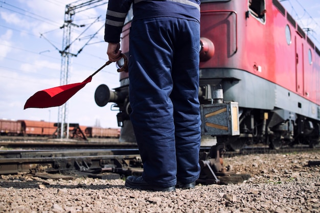 Railroad worker or switchman with red flag standing by the rail tracks as train passing by at the station.