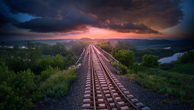 Railway and bridge over the river on a background of sunset and storm clouds. Aerial view. Beautiful summer evening landscape.