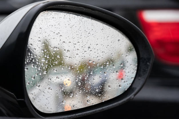 後ろの車の焦点がぼけた反射を伴う車のバックミラーの雨滴