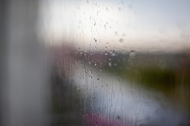 曇り灰色の空と都市の建物の背景の窓から雨の日。概念。ガラス窓の後ろの夜の街並みと水滴が滴り落ちる。