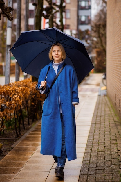 Concetto di tempo piovoso una donna in un cappotto blu con un ombrello blu su una strada cittadina donna quaranta anni più