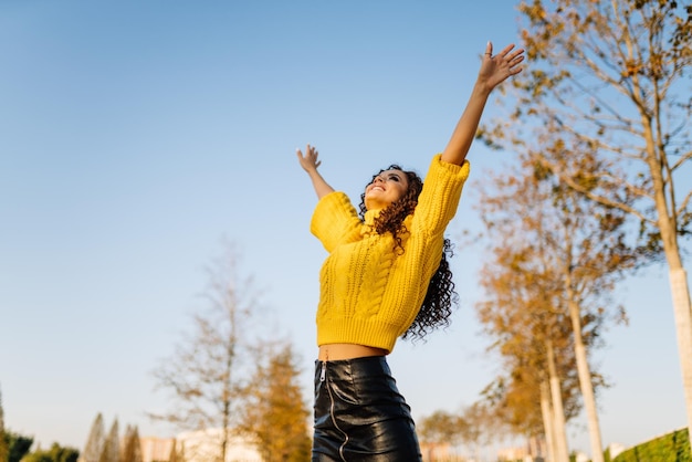 Raising her hands up and wide of them spread curly girl with dark hair happily looks into the sky in