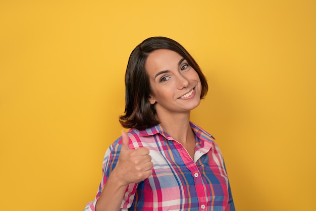Raising thumb up gesture with hand smile beautiful woman dressed in a plaid shirt and dark hair on yellow wall