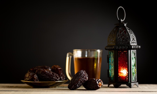 Photo ramadan food and drinks concept ramadan lantern with arabian lamp wood rosary tea dates fruit and lighting on a wooden table with dark background