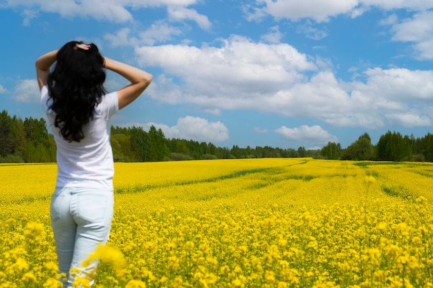 Rapeseed field with yellow flowers, blue sky with clouds and darkhaired beautiful brunette girl, view from the back. copy space