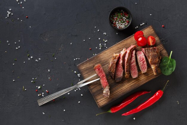 Rare rib eye steaks with herbs and spices on wooden board with meat fork, tomato and chilli at dark background with copy space, top view