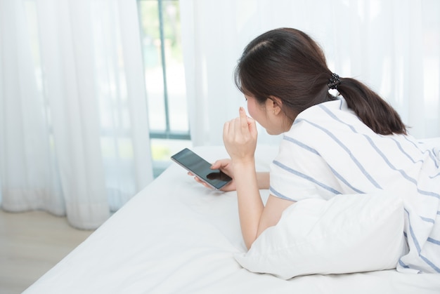 Rare view or back side of asian woman lay down on bed, holding smart phone and looking up thing something