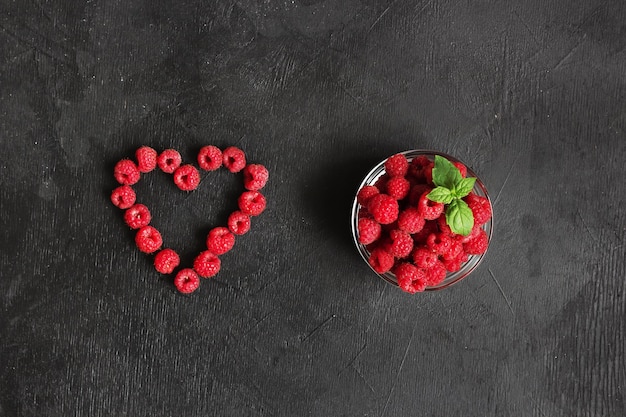 Photo the raspberies heart and fresh raspberries in the clear glass bowl on the black background