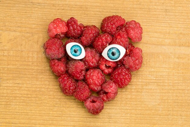 Raspberries in a heart shape with toy blue eyes on a wooden table