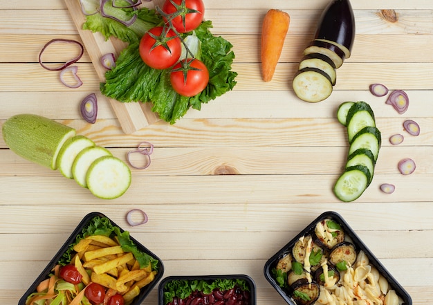 Rauwe groenten rond houten tafel. Zwarte voedselcontainers met lunch en diner. Kopieer ruimte