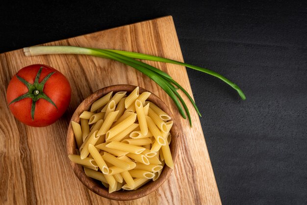 Rauwe pasta in houten kom met kaas, tomaat en groene ui op zwarte achtergrond.