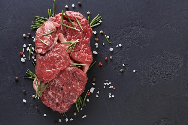 Rauwe rib eye steak met kruiden en groenten. Ingrediënten voor restaurantmaaltijd. Vers vlees, zout, rozemarijn, tijm, chili, kerstomaten, knoflook op zwarte steen. Voedselachtergrond, kopieerruimte, bovenaanzicht