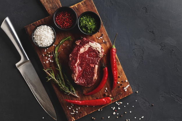 Rauwe rib eye steak met kruiden en mes, close-up. Kokende ingrediënten voor restaurantschotel. Vers vlees, specerijen en chili op houten bureau, bovenaanzicht