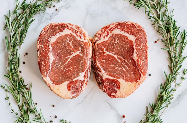 Rauwe twee ribeye steaks in het midden van een kerstkrans van rozemarijn tijm cherrytomaatjes en paprika's op een wit marmeren oppervlak kerstdiner concept voor twee