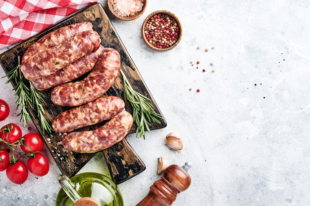 Rauwe worstjes of braadworst op snijplank met kruiden en ingrediënten om te koken. Bovenaanzicht met kopieerruimte op lichtgrijze stenen tafel.