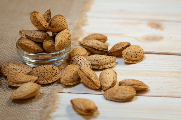 Raw almonds in the skin lies in a glass bowl and scattered on a wooden background, close-up