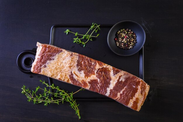 Raw bacon strips with fresh spices on a black ccutting board.