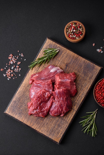 Raw beef cut into several pieces on a wooden cutting board on a dark concrete background