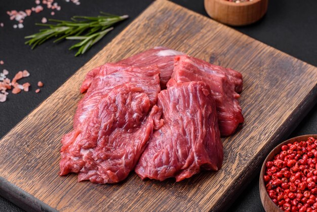 Raw beef cut into several pieces on a wooden cutting board on a dark concrete background