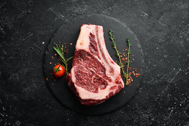 Raw beef ribeye steak on the bone with thyme rosemary and spices Top view Flat lay top view on black stone cutting table