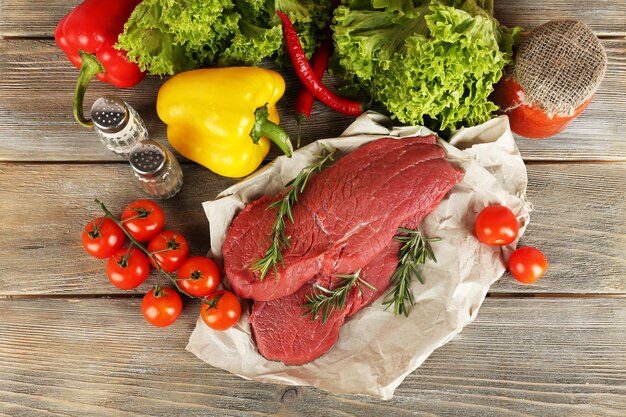 Raw beef steak on paper with vegetables and greens on wooden background