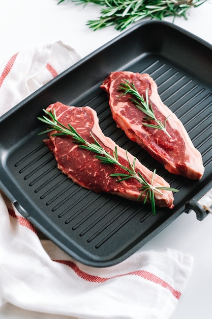 Raw beef steak with rosemary in black grill pan on the table.