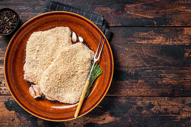 Raw Breaded German Wiener schnitzel on a rustic plate with herbs. Dark wooden background. Top view. Copy space.