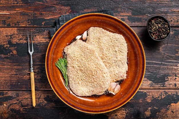 Raw Breaded German Wiener schnitzel on a rustic plate with herbs. Dark wooden background. Top view.