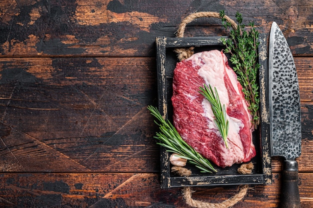 Raw brisket beef meat cut on a wooden tray with knife. Dark wooden background. Top view. Copy space.