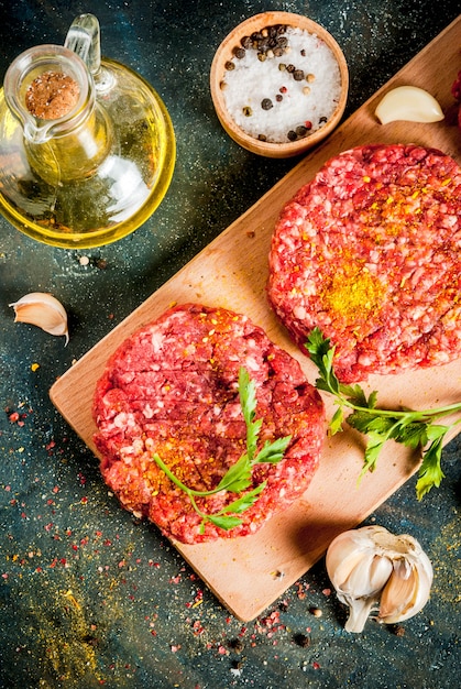 Raw burger cutlets with salt, pepper, oil, herbs and spice, on dark table, copyspace top view