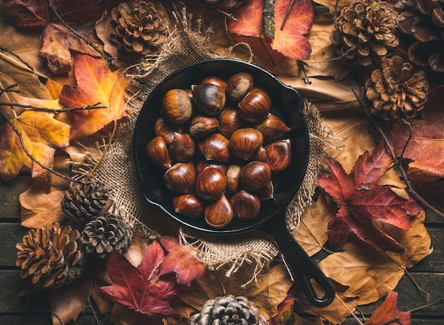 Foto castagne crude in una piccola padella di ferro su un tavolo con foglie autunnali e pigne