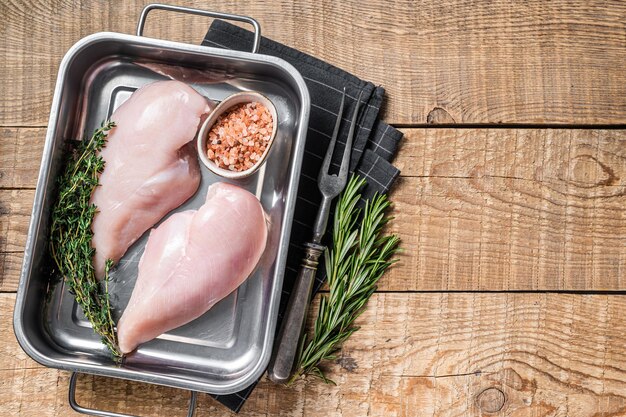 Raw chicken breast fillet in kitchen tray with thyme and rosemary. Wooden background. Top view. Copy space.