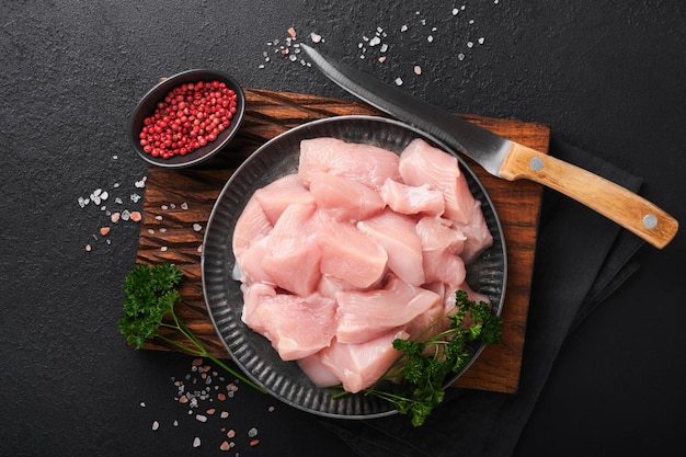 Raw chicken breast sliced or cut pieces on wooden cutting board\
with herbs and spices on dark slate stone concrete background raw\
chicken meat top view with copy space mock up