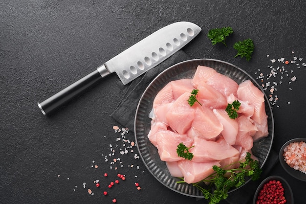 Raw chicken breast sliced or cut pieces on wooden cutting board\
with herbs and spices on dark slate stone concrete background raw\
chicken meat top view with copy space mock up
