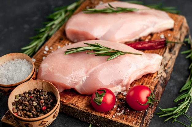 Raw chicken fillet on a cutting board with rosemary, spices, tomatoes and pepper on a stone background