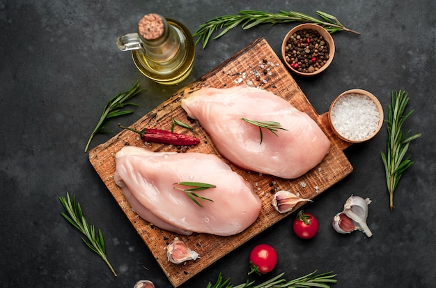 Filetto di pollo crudo su un tagliere con rosmarino, spezie, pomodori e pepe su uno sfondo di pietra