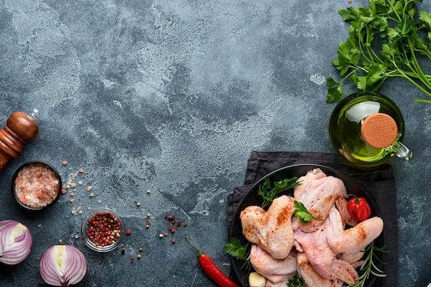 Raw chicken wings in metal pan or bowl with spices and ingredients for cooking on dark grey slate, stone or concrete background. Raw meat with spices at black table. Top view. Mock up.